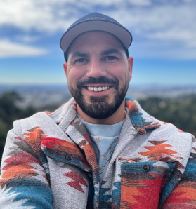 Photo head-and-shoulders shot of 30-year-old bearded man smiling broadly and wearing a zip-up jacket bearing Native American art design like a Navajo rug